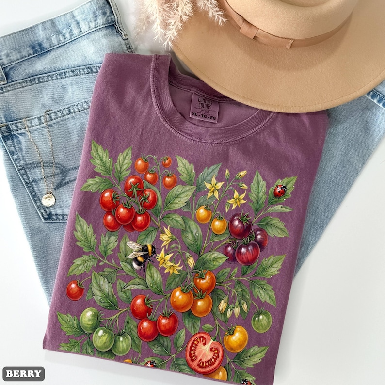 Op de afbeelding: Een bessenkleurig T-shirt met een gedetailleerde illustratie van een tomatenplant met rode, oranje en groene tomaten, gele bloemen, een hommel en een lieveheersbeestje. Het shirt is opgevouwen op een spijkerbroek, met een ketting en een beige hoed.