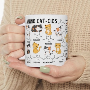 Może przedstawiać: Biały kubek ceramiczny z kolorową ilustracją kot&oacute;w i wzor&oacute;w chemicznych. Na kubku nadrukowany jest tekst "AMINO CAT-CIDS". Wzory chemiczne są oznaczone nazwami związanymi z kotami, takimi jak "MEOWLINE" i "PURRLINE".