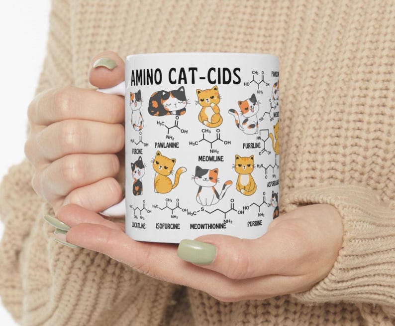 Może przedstawiać: Biały kubek ceramiczny z kolorową ilustracją kot&oacute;w i wzor&oacute;w chemicznych. Na kubku nadrukowany jest tekst "AMINO CAT-CIDS". Wzory chemiczne są oznaczone nazwami związanymi z kotami, takimi jak "MEOWLINE" i "PURRLINE".