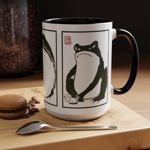 May include: White ceramic mug with a black interior and handle. The mug features a black and white illustration of a frog with a bowl, and a fish. The mug is sitting on a wooden cutting board with a spoon and a macaron.