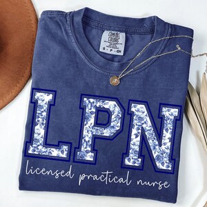 May include: A navy blue t-shirt with the letters "LPN" in a floral pattern, outlined in dark blue. The words "licensed practical nurse" are written below the letters in a cursive font. A gold necklace is draped across the shirt.