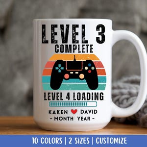 May include: White ceramic mug with a retro-style design featuring a video game controller and the text "Level 3 Complete Level 4 Loading". The mug also includes a progress bar with a heart icon and the text "Kaken - Month Year - David".