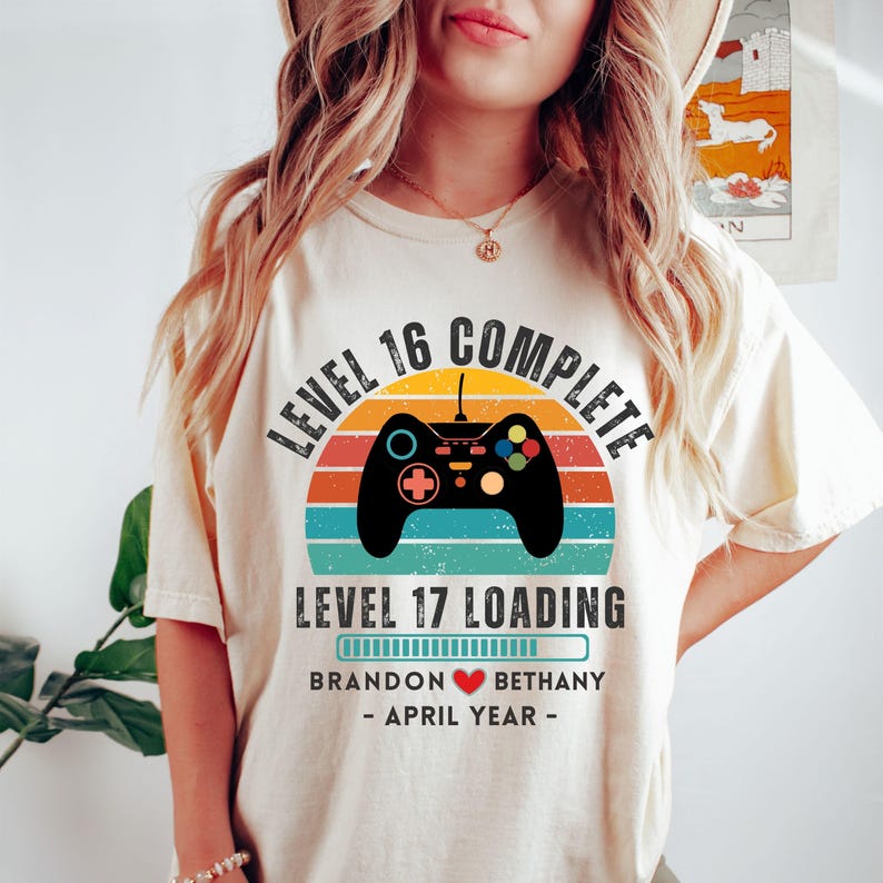 May include: A white t-shirt with a retro-style graphic that says "Level 16 Complete" and "Level 17 Loading" with a progress bar. The graphic also includes a video game controller and a sunset background. The text "Brandon Bethany - April Year -" is also included on the shirt.