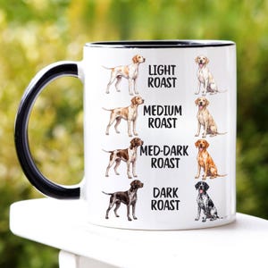Könnte beinhalten: Ein weißer Keramikbecher mit schwarzem Henkel und Innenseite, bedruckt mit Hundeillustrationen in vier Farbvarianten sowie dem Text Light Roast, Medium Roast, Med-Dark Roast und Dark Roast, die verschiedene Fellmuster zeigen.