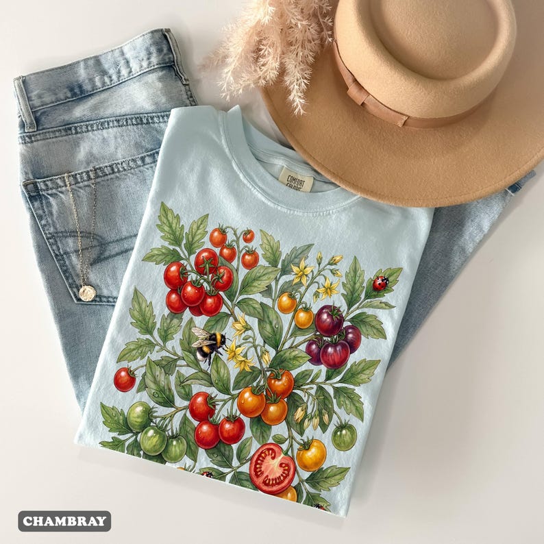 Op de afbeelding: Een lichtblauw t-shirt met een tomatenplantontwerp, inclusief rode, oranje en groene tomaten, een hommel en een lieveheersbeestje. Het shirt wordt gecombineerd met een lichtblauwe jeans en een beige hoed.