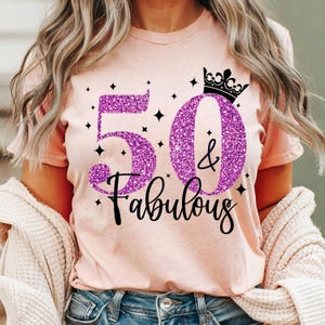 May include: A light peach-colored t-shirt featuring a graphic print with the number 50 in purple glitter, a black crown icon above the zero, and the word Fabulous in black script. Small black star accents surround the text and numbers.