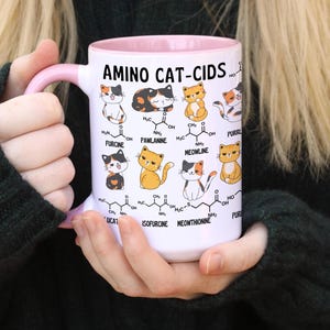 Może przedstawiać: Biały kubek ceramiczny z r&oacute;żowym rantem i uchwytem. Kubek przedstawia humorystyczną ilustrację kot&oacute;w ze strukturami chemicznymi i tekstem "AMINO CAT-CIDS".