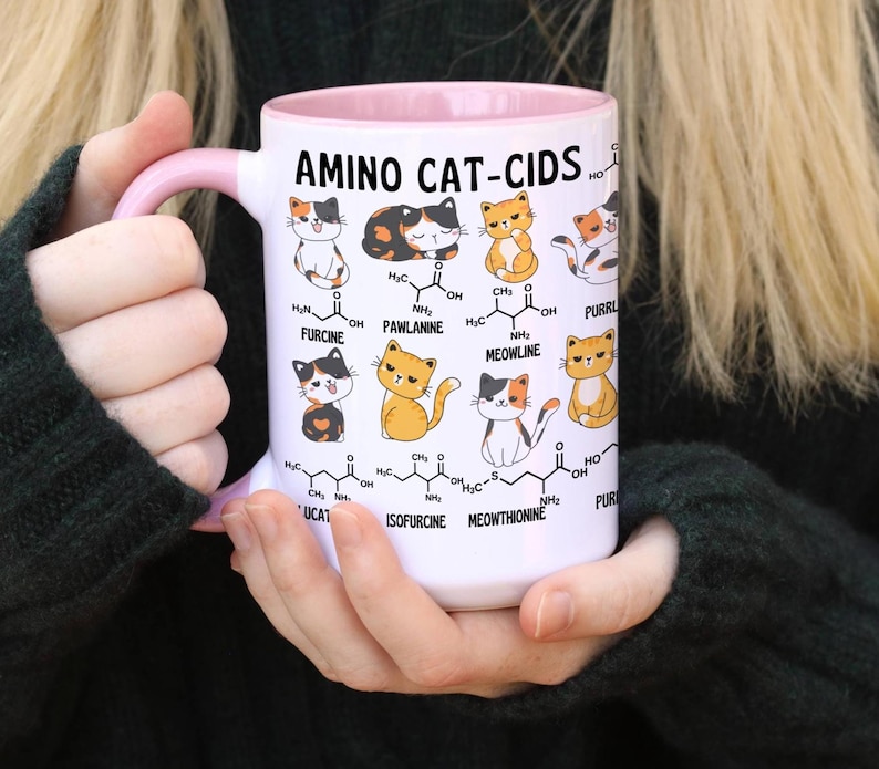 Może przedstawiać: Biały kubek ceramiczny z r&oacute;żowym rantem i uchwytem. Kubek przedstawia humorystyczną ilustrację kot&oacute;w ze strukturami chemicznymi i tekstem "AMINO CAT-CIDS".
