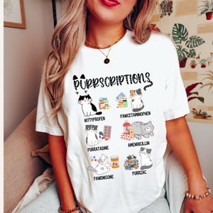 Könnte beinhalten: Weißes T-Shirt mit dem Text "Purrscriptions" und Illustrationen von Katzen und Medikamentenflaschen. Das Shirt zeigt verschiedene Medikamentennamen zum Thema Katze wie "Kittyprofen" und "Purrzac".