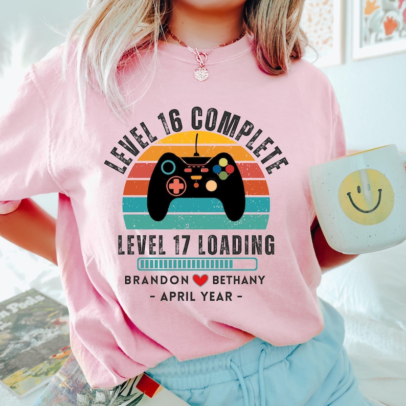 May include: Pink t-shirt with a retro video game controller graphic and the text "Level 16 Complete" and "Level 17 Loading". The graphic also includes a progress bar with the text "Brandon Bethany - April Year -"