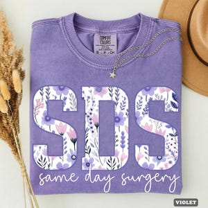 May include: A violet t-shirt with the letters "SDS" in a floral design, with the words "same day surgery" below. A silver necklace with a star pendant and a brown hat are also visible.