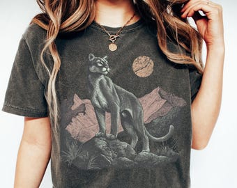 Könnte beinhalten: Dunkelgraues T-Shirt mit einer Illustration eines Pumas. Das Design zeigt einen Puma auf einem Felsen, mit Bergen und einer Sonne im Hintergrund. Das T-Shirt hat einen Vintage-Waschstil.