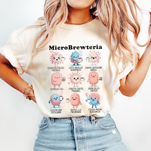 Może przedstawiać: Kremowa koszulka z napisem "MicroBrewteria" nad kreskówkowymi ilustracjami mikrobów trzymających filiżanki kawy. Mikrobi są w różnych kolorach, w tym różowym, niebieskim i pomarańczowym. Koszulka ma okrągły dekolt i krótkie rękawy.