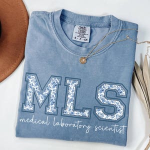 Op de afbeelding: Een lichtblauw t-shirt met de letters "MLS" in een varsity-stijl, gevuld met een blauw bloemenpatroon. De woorden "medical laboratory scientist" staan onder de letters in een scriptlettertype. Een gouden ketting is over het shirt gedrapeerd.