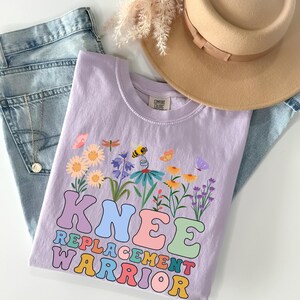 Könnte beinhalten: Ein helllila T-Shirt mit floralem Design und dem Schriftzug "KNEE REPLACEMENT WARRIOR" in bunten Buchstaben. Das Shirt wird mit einer blauen Jeans und einem beigen Hut präsentiert.