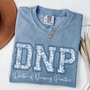 Può includere: Una maglietta blu Comfort Colors con le lettere "DNP" in un motivo floreale. Il testo "Doctor of Nursing Practice" è sotto. Una collana d'oro con un ciondolo è sulla maglietta.