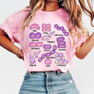 May include: Pale pink t-shirt displaying a colourful illustration of different bacteria, such as Vibrio, Tetrad, and Streptococci. The design includes the names of each bacteria type. The t-shirt is a light pink colour.
