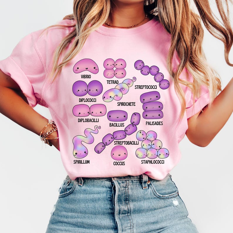 May include: Pale pink t-shirt displaying a colourful illustration of different bacteria, such as Vibrio, Tetrad, and Streptococci. The design includes the names of each bacteria type. The t-shirt is a light pink colour.