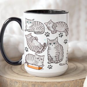 May include: White ceramic mug with a black handle and rim. The mug features a repeating pattern of gray and white striped cats in various poses. The cats have black eyes and noses. The background is white with gray paw prints.