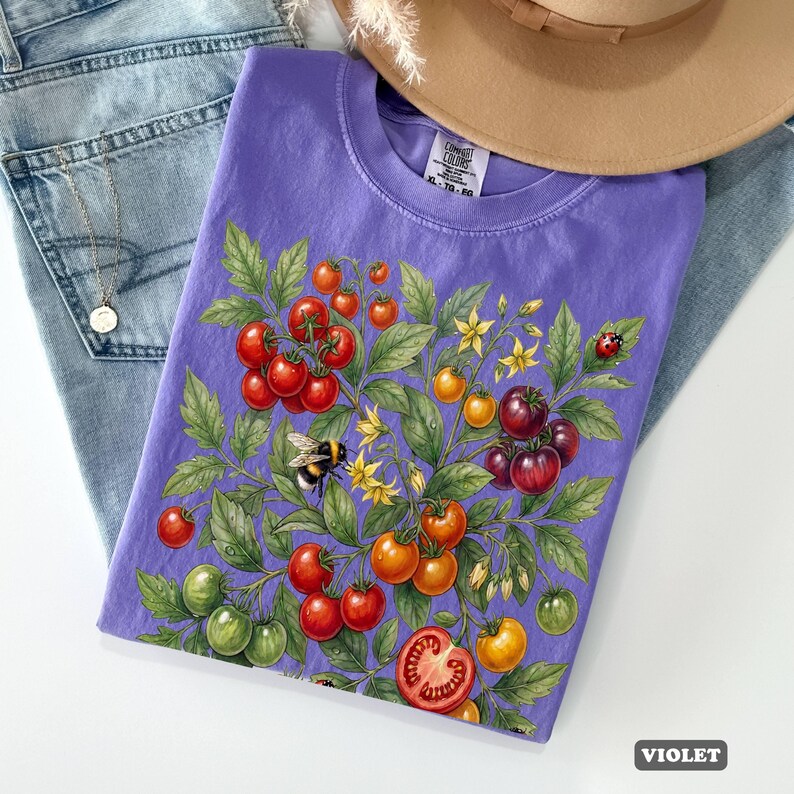 Op de afbeelding: Een violetkleurig T-shirt met een gedetailleerde illustratie van een tomatenplant met rijpe rode, oranje en groene tomaten, gele bloemen, een lieveheersbeestje en een hommel. Het shirt ligt op een blauwe jeans en een bruine hoed.