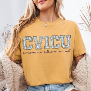 May include: Mustard yellow t-shirt with the text "CVICU" in blue floral letters. Below the letters is the text "cardiovascular intensive care unit" in a cursive font. The shirt has short sleeves and a crew neck.