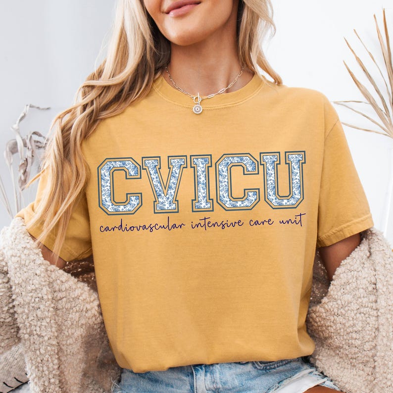 May include: Mustard yellow t-shirt with the text "CVICU" in blue floral letters. Below the letters is the text "cardiovascular intensive care unit" in a cursive font. The shirt has short sleeves and a crew neck.