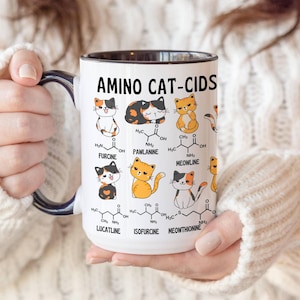 Może przedstawiać: Biały kubek ceramiczny z czarnym rantem i uchwytem. Kubek przedstawia humorystyczną ilustrację kot&oacute;w ze wzorami chemicznymi i tekstem "Amino Cat-Cids".