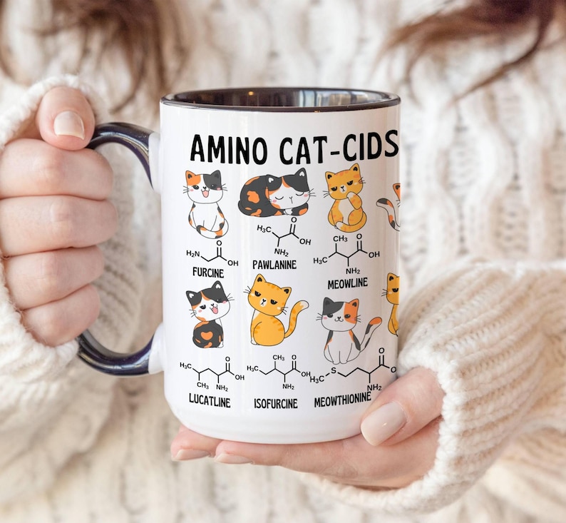 Może przedstawiać: Biały kubek ceramiczny z czarnym rantem i uchwytem. Kubek przedstawia humorystyczną ilustrację kot&oacute;w ze wzorami chemicznymi i tekstem "Amino Cat-Cids".