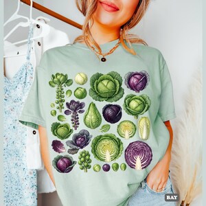 Könnte beinhalten: Hellgrünes T-Shirt mit Illustrationen verschiedener Kohlsorten in Grün-, Lila- und Gelbtönen. Das Design umfasst ganze und geschnittene Kohlköpfe sowie andere verwandte Pflanzen. Das Wort "BAY" ist unten rechts sichtbar.