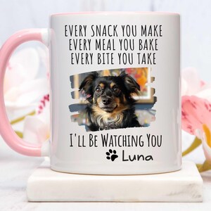 May include: White ceramic coffee mug with a pink handle. The mug features a black and white photo of a dog with the text "EVERY SNACK YOU MAKE EVERY MEAL YOU BAKE EVERY BITE YOU TAKE I'LL BE WATCHING YOU Luna".