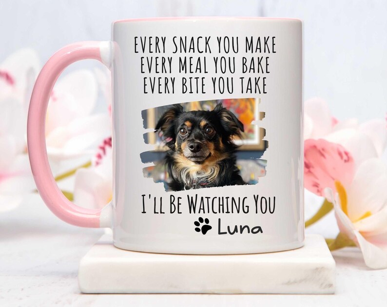 May include: White ceramic coffee mug with a pink handle. The mug features a black and white photo of a dog with the text "EVERY SNACK YOU MAKE EVERY MEAL YOU BAKE EVERY BITE YOU TAKE I'LL BE WATCHING YOU Luna".