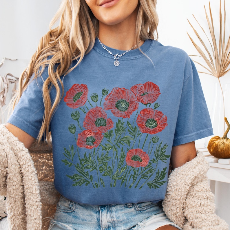 May include: A blue t-shirt featuring a red poppy flower design. The graphic includes detailed red poppies with green leaves and stems. The shirt is a comfortable, casual style.