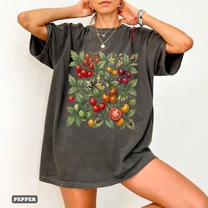 Op de afbeelding: Donkergrijs t-shirt met een kleurrijk tomatenplantontwerp. Het ontwerp bevat rode, oranje, gele en groene tomaten, groene bladeren, een bij en lieveheersbeestjes. Het woord "PEPPER" staat linksonder.