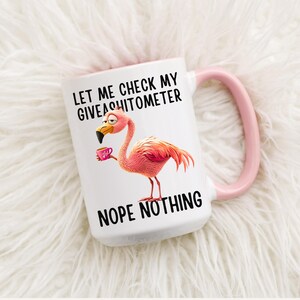 Könnte beinhalten: Eine weiße Keramiktasse mit rosafarbenem Rand und Henkel. Die Tasse zeigt einen rosa Flamingo mit einer Tasse Kaffee und dem Text "Let me check my giveashitometer Nope nothing".