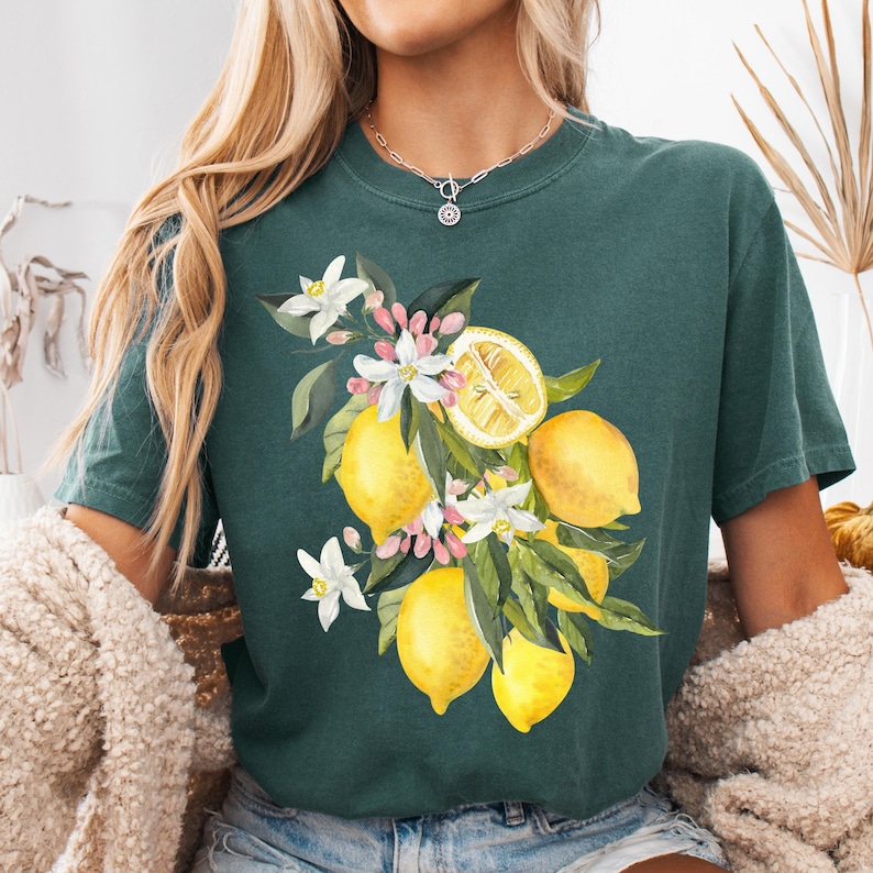 May include: Dark teal t-shirt featuring a watercolor illustration of lemons, white flowers, and green leaves. The lemons are yellow, and the flowers have pink accents. The shirt has a classic crew neck and short sleeves.