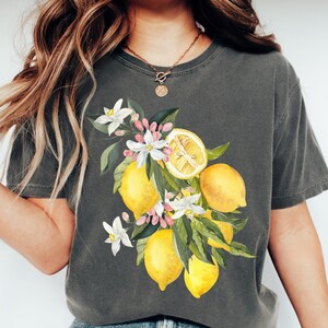 May include: Dark gray t-shirt featuring a watercolor illustration of lemons, white flowers, and green leaves. The design includes whole and sliced lemons, creating a botanical theme. The shirt is a comfortable, casual style.