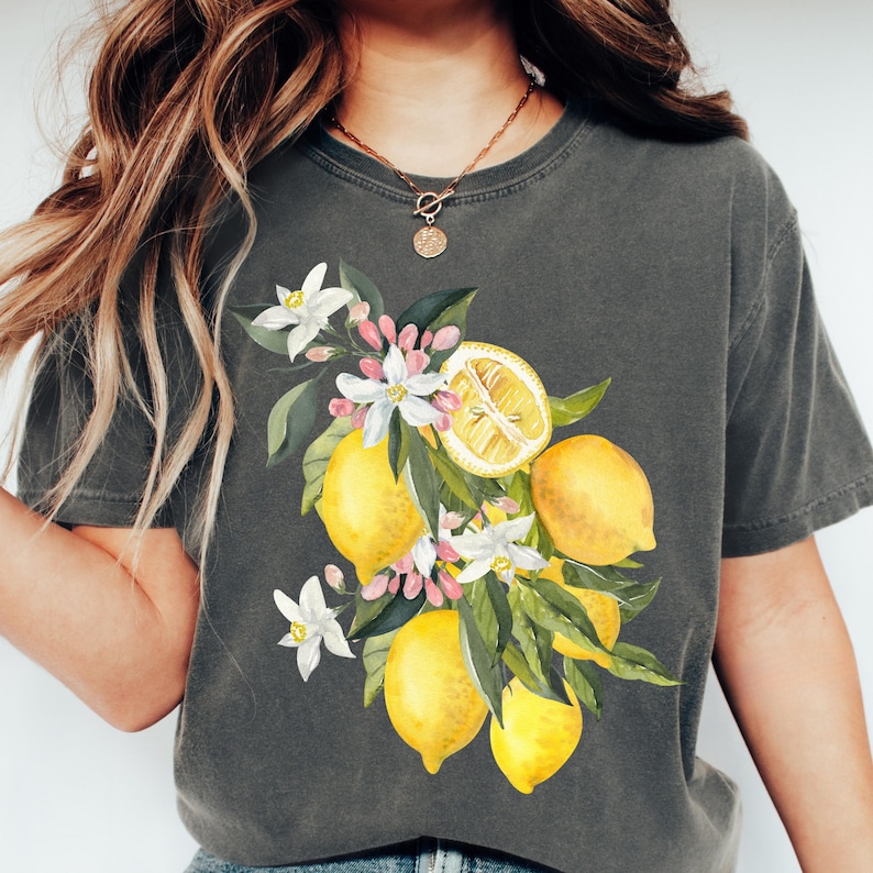 May include: Dark gray t-shirt featuring a watercolor illustration of lemons, white flowers, and green leaves. The design includes whole and sliced lemons, creating a botanical theme. The shirt is a comfortable, casual style.