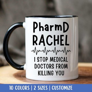 Puede incluir: Taza de café blanca con borde y asa negros. La taza tiene texto negro que dice "PharmD RACHEL I STOP MEDICAL DOCTORS FROM KILLING YOU" con una línea negra de ECG sobre el texto.