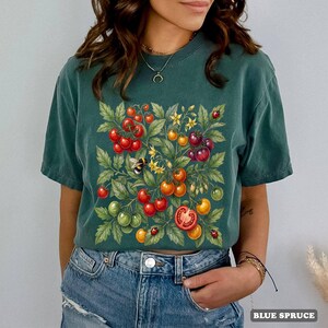 Op de afbeelding: Een teal-kleurig T-shirt met een gedetailleerde illustratie van tomaten, bladeren, een bij en lieveheersbeestjes. De tomaten zijn rood, oranje en groen. De tekst "BLUE SPRUCE" staat rechtsonder.
