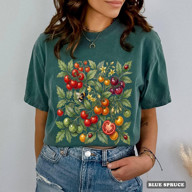 Op de afbeelding: Een teal-kleurig T-shirt met een gedetailleerde illustratie van tomaten, bladeren, een bij en lieveheersbeestjes. De tomaten zijn rood, oranje en groen. De tekst "BLUE SPRUCE" staat rechtsonder.