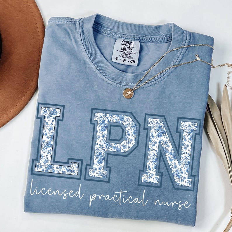May include: A blue Comfort Colors t-shirt with the letters "LPN" in a floral design. The text "licensed practical nurse" is below the letters. A gold necklace and a brown hat are also visible.