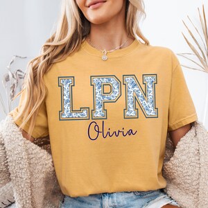 May include: Mustard yellow t-shirt with the letters "LPN" in a blue floral pattern, outlined in a darker blue. The name "Olivia" is written in cursive below the letters. The shirt is a casual style.