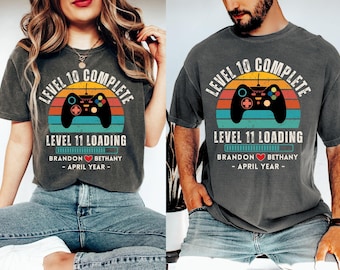 May include: Two gray t-shirts with a retro video game design. The design includes a controller, a sunset, and the text "LEVEL 10 COMPLETE" and "LEVEL 11 LOADING". The shirts also display names and a year.