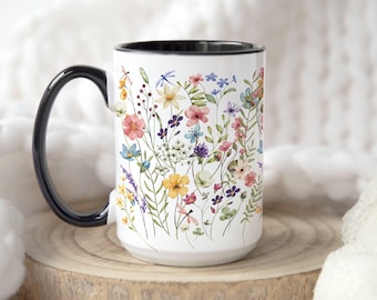 Può includere: Tazza in ceramica bianca con manico e bordo neri, ornata da un colorato motivo floreale. I fiori includono fiori rosa, gialli, blu e viola, con foglie e steli verdi. La tazza è appoggiata su una superficie di legno.