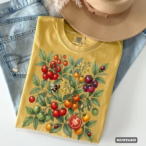 Op de afbeelding: Mosterdkleurig T-shirt met een ontwerp van een tomatenplant. De afbeelding toont rode, oranje en groene tomaten, een bij en lieveheersbeestjes. Het shirt wordt gecombineerd met een blauwe jeans en een bruine hoed. De tekst "COMFORT COLORS" is zichtbaar op het label.