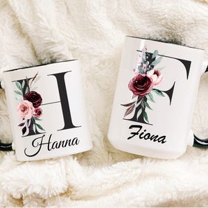 Puede incluir: Dos tazas de cerámica blanca con bordes y asas negros. Las tazas presentan una gran letra negra, un diseño floral y un nombre. La taza de la izquierda tiene la letra "H" y el nombre "Hanna". La taza de la derecha tiene la letra "F" y el nombre "Fiona".