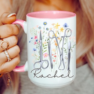 Könnte beinhalten: Weiße Keramiktasse mit rosafarbenem Rand und Henkel. Die Tasse zeigt ein Blumenmuster mit chirurgischen Instrumenten und den Namen "Rachel" in schwarzer Schrift.