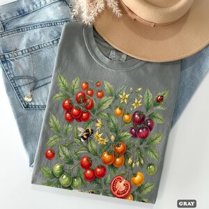 Op de afbeelding: Grijze t-shirt met een aquarel illustratie van een tomatenplant met rode, oranje en groene tomaten, gele bloemen, een hommel en een lieveheersbeestje. Het shirt wordt gecombineerd met een blauwe jeans, een ketting en een beige hoed.