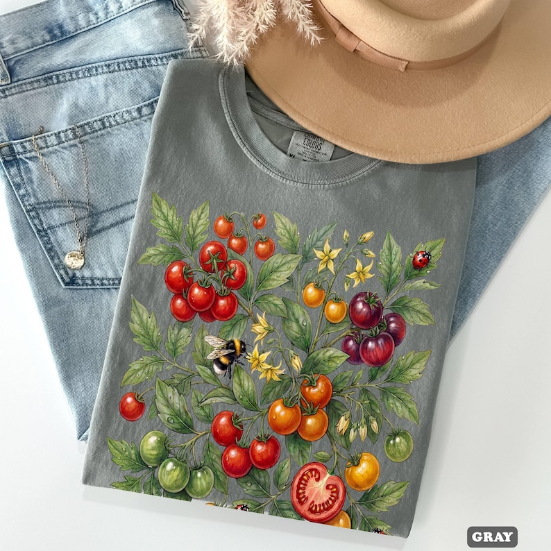 Op de afbeelding: Grijze t-shirt met een aquarel illustratie van een tomatenplant met rode, oranje en groene tomaten, gele bloemen, een hommel en een lieveheersbeestje. Het shirt wordt gecombineerd met een blauwe jeans, een ketting en een beige hoed.