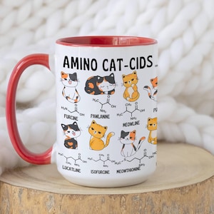Może przedstawiać: Biały kubek ceramiczny z czerwonym rantem i uchwytem. Kubek przedstawia kresk&oacute;wkową ilustrację kot&oacute;w ze wzorami chemicznymi i tekstem "AMINO CAT-CIDS".